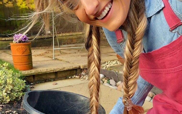 Young woman with braided hair smiling whilst filling a black bucket with peat-free compost in a garden setting.