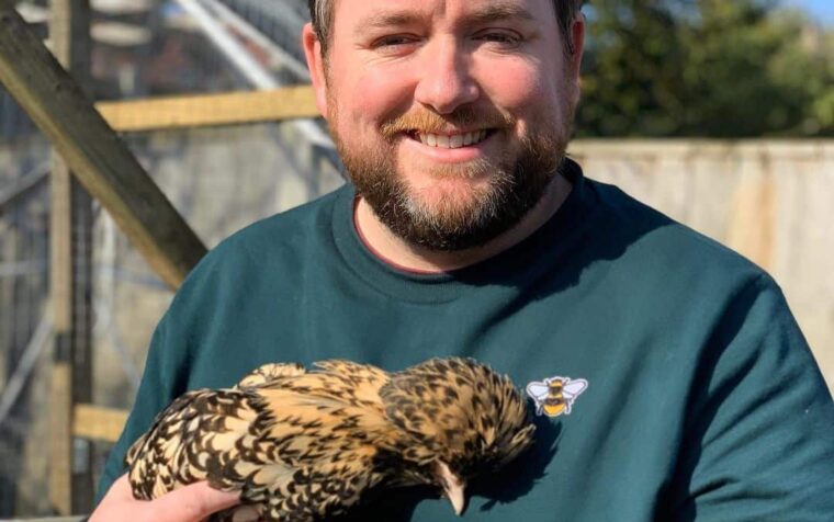 Man holding a bird of prey outdoors, wearing green shirt with bee logo, demonstrating wildlife interaction in garden setting.