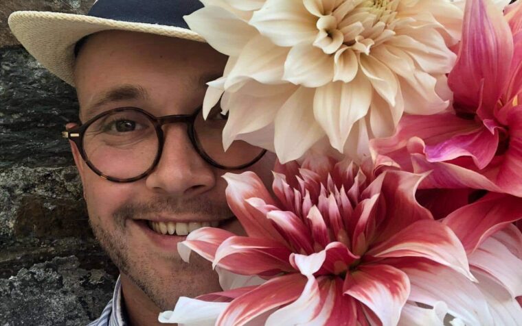 Man wearing straw hat and glasses holding large peach and pink dahlias against stone wall backdrop.