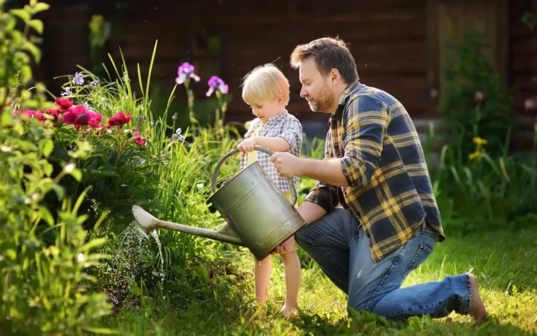 Adult and young child watering red flowers in a garden with a watering can, surrounded by green plants and purple blooms.