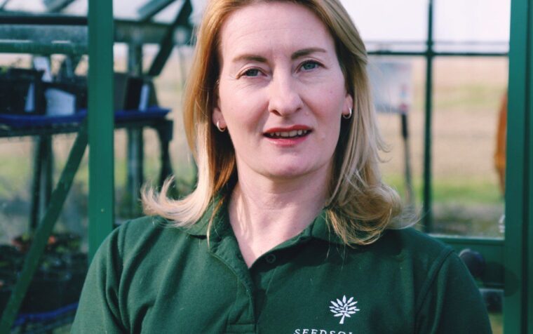 Woman in green Seedsology polo shirt standing in greenhouse, smiling at camera with fields visible behind.