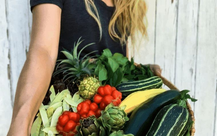 Woman holding woven basket filled with fresh vegetables including artichokes, tomatoes, courgettes, and pineapple against white wooden background.