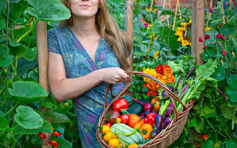 Woman holding wicker basket of harvested vegetables and flowers in productive garden with trellises and flowering plants.