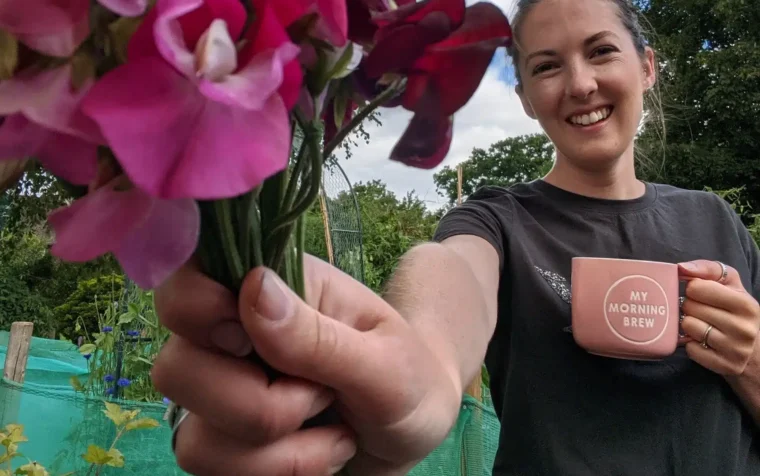 Woman holding vibrant magenta sweet pea flowers and a mug in a garden setting.