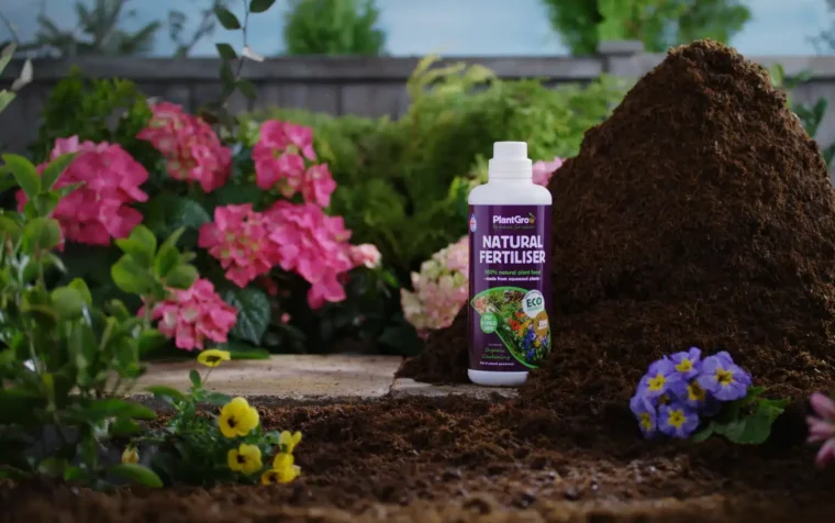 PlantGrow natural fertiliser bottle displayed in garden setting surrounded by blooming flowers and soil.