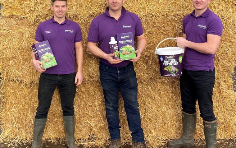 Three PlantGrow team members in purple branded polos holding slug and snail barrier, liquid feed, and natural fertiliser products against straw backdrop.