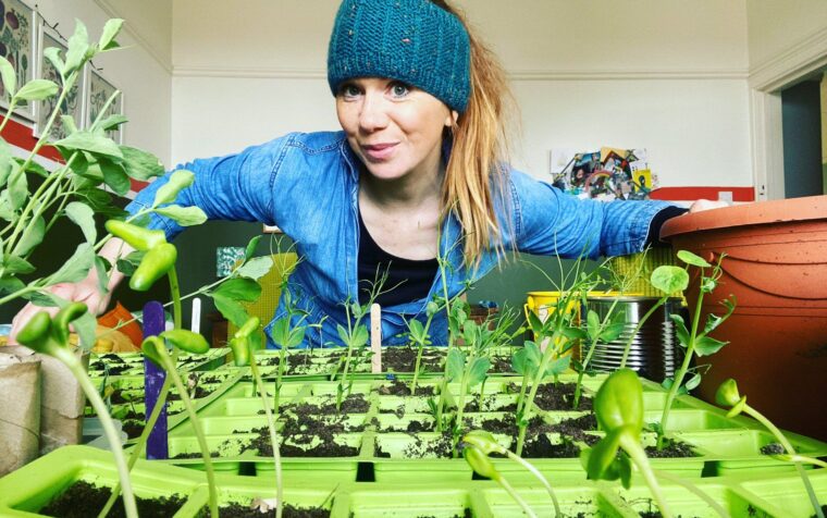 Woman in blue denim shirt and headband tending seedlings in bright green plant trays indoors.