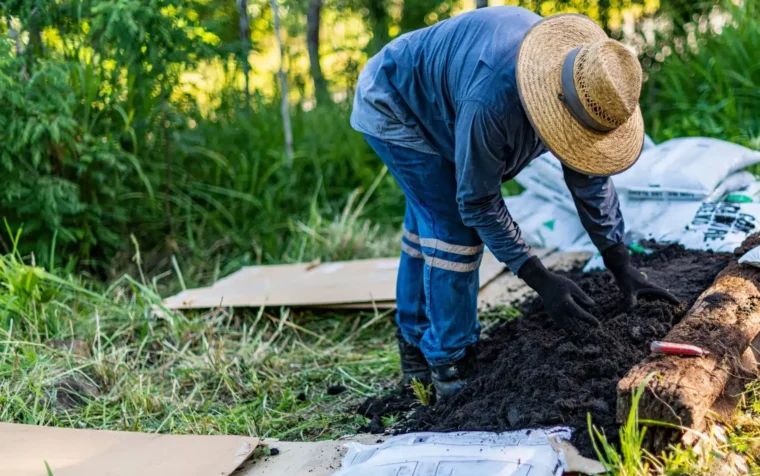 Gardener in straw hat using no dig method, layering compost and soil over cardboard in garden bed.