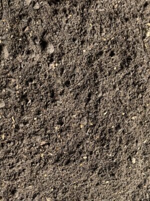 Close-up of PlantGrow Raised Bed Mix: dark, loose, and slightly moist soil with small pebbles and organic matter visible throughout the surface.