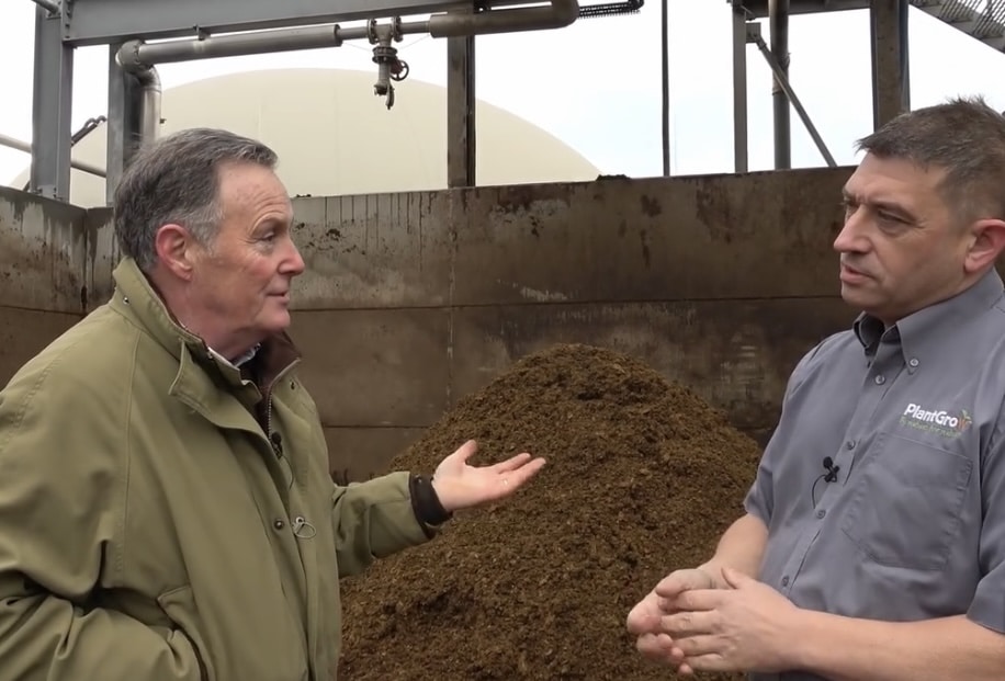 Two men are having a conversation in front of a large pile of organic material in an industrial setting. One man is gesturing with his hand, while the other listens with his hands clasped. Both are wearing jackets.