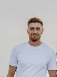A man with short light brown hair and a beard is wearing a plain white t-shirt and smiling at the camera against a light, neutral background.