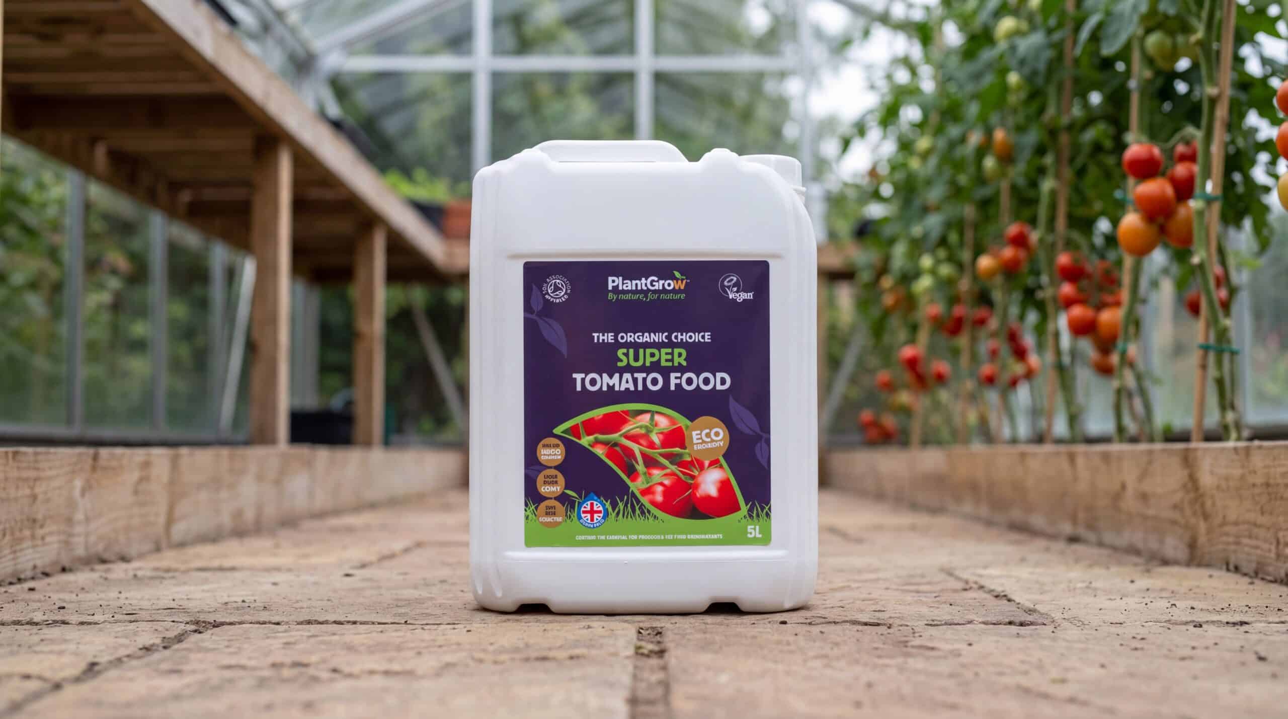PlantGrow Super Tomato Food, a 5-litre bottle of organic liquid fertiliser positioned on a greenhouse path with tomato plants growing behind.